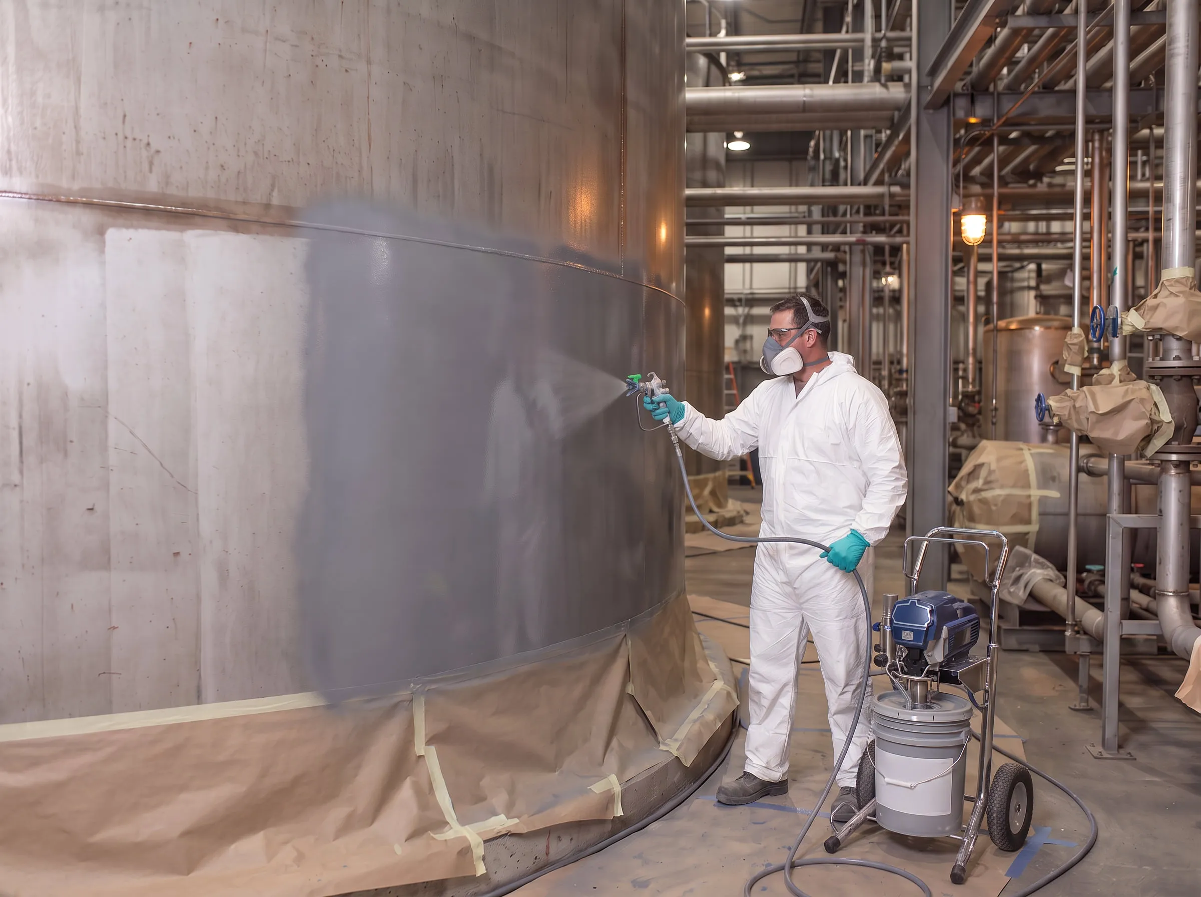 Worker in protective coveralls and a respirator spray-applying a grey coating to the exterior of a large steel tank in an industrial facility.